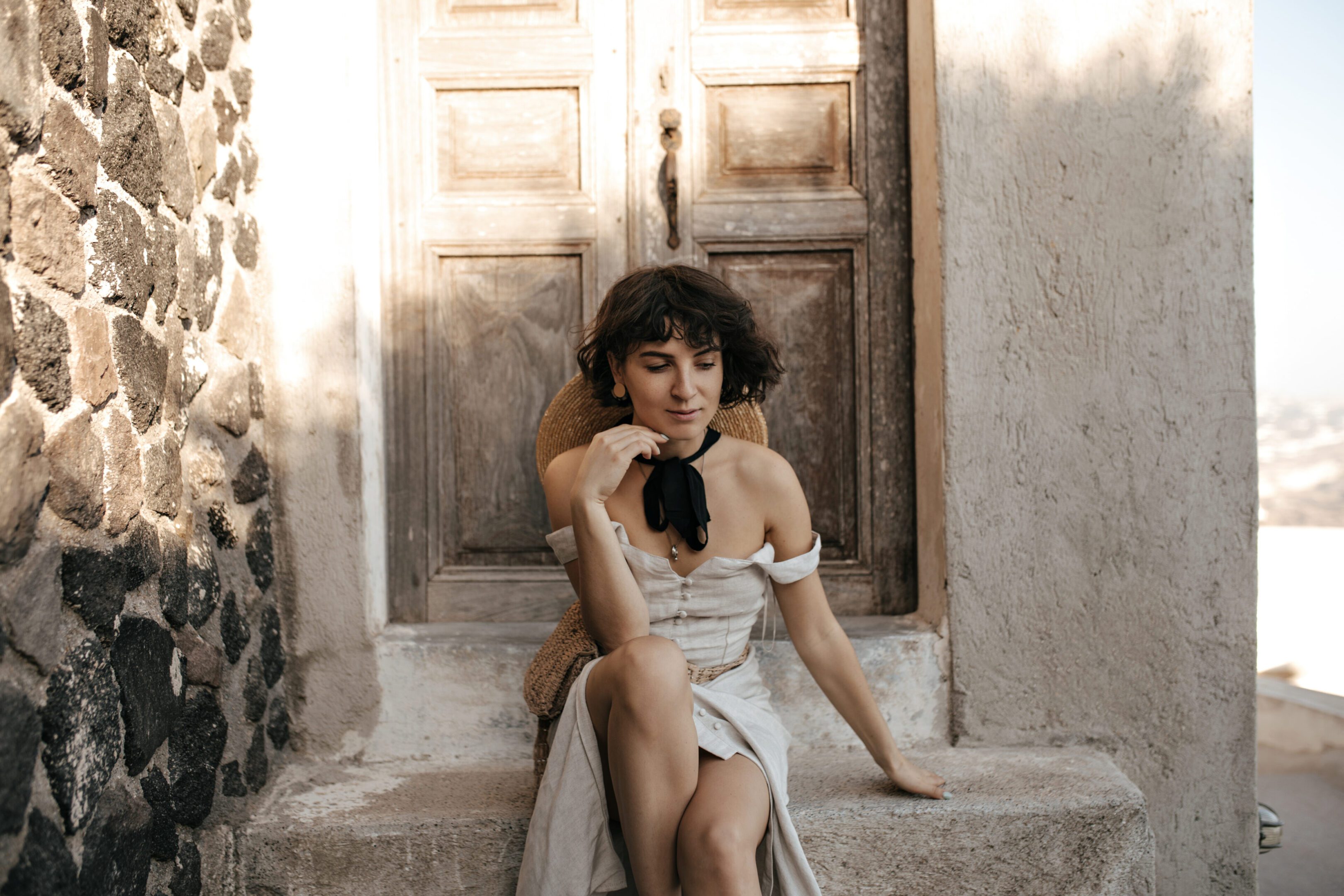 A woman in a white dress sits on stone steps in front of a wooden door.