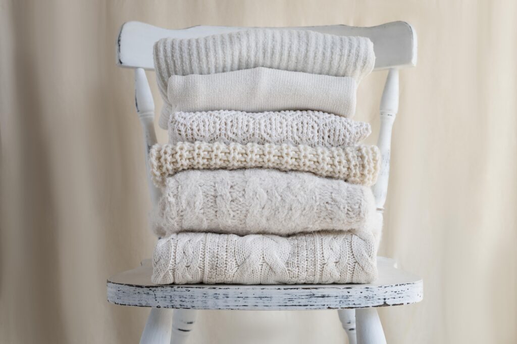 Stack of cozy, knit sweaters in neutral tones on a chair.