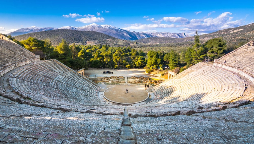 Ancient open-air amphitheater with scenic mountain backdrop under a blue sky.