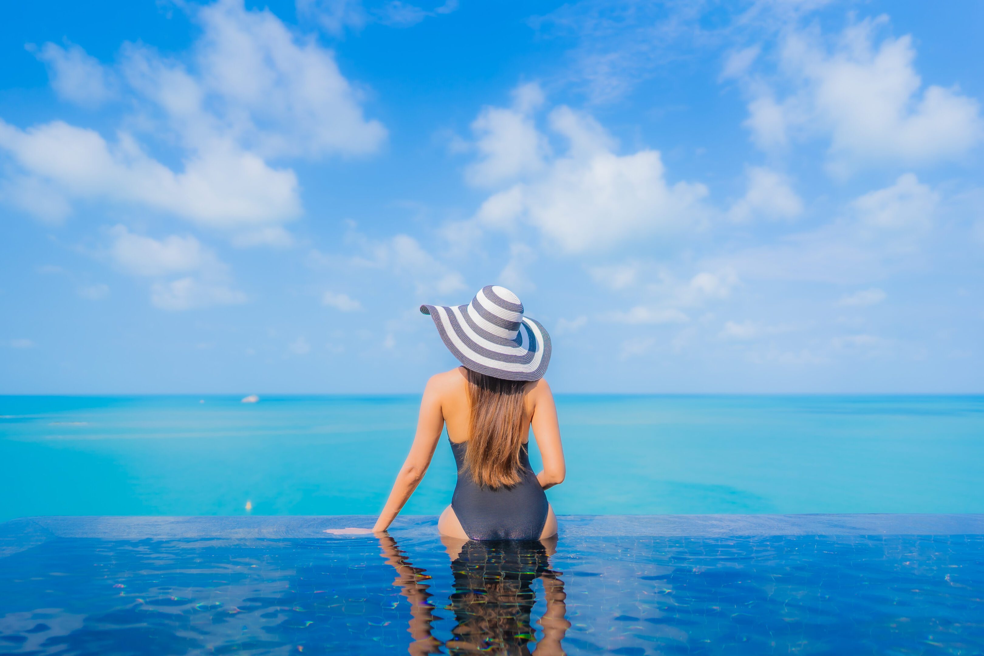 Woman relaxing by an oceanfront pool with a wide sea view, enjoying a peaceful wellness moment under clear blue skies.