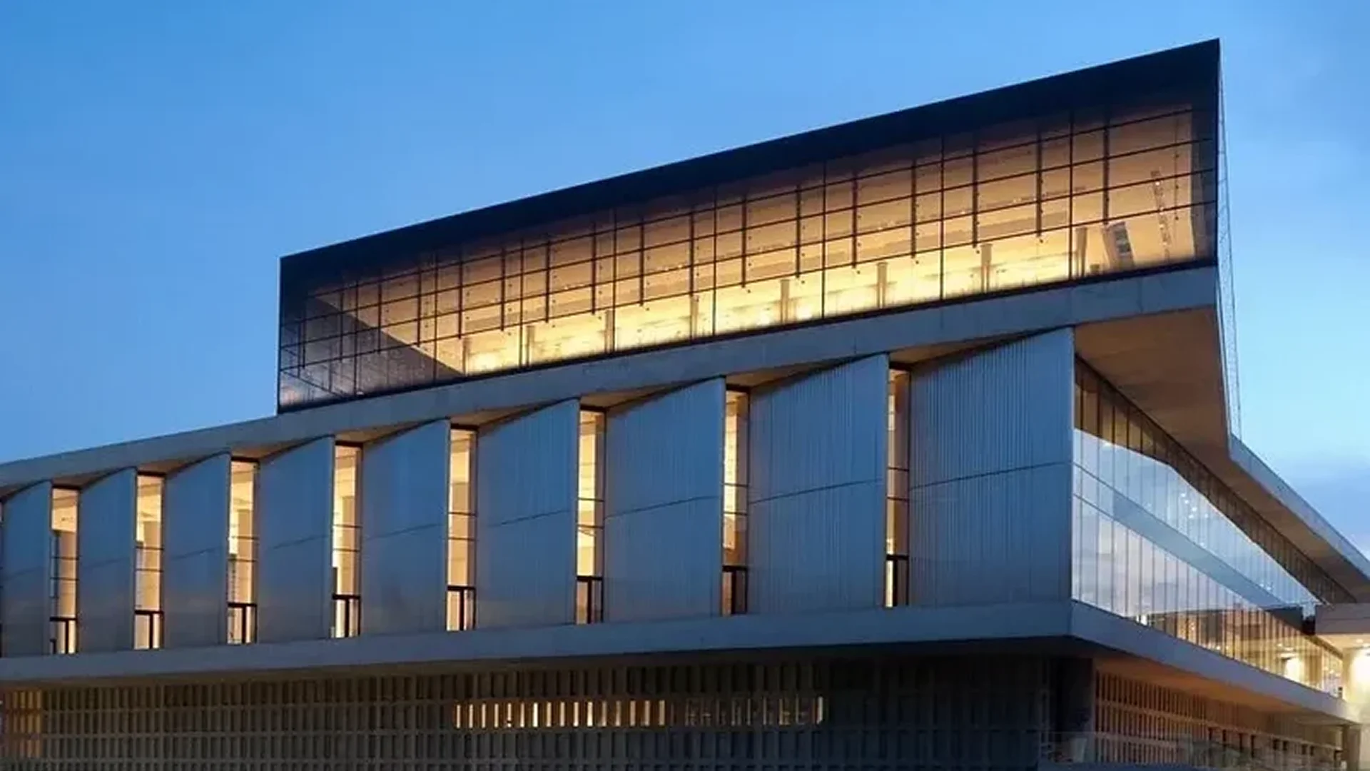 Modern building with tall pillars and illuminated upper floor at dusk.