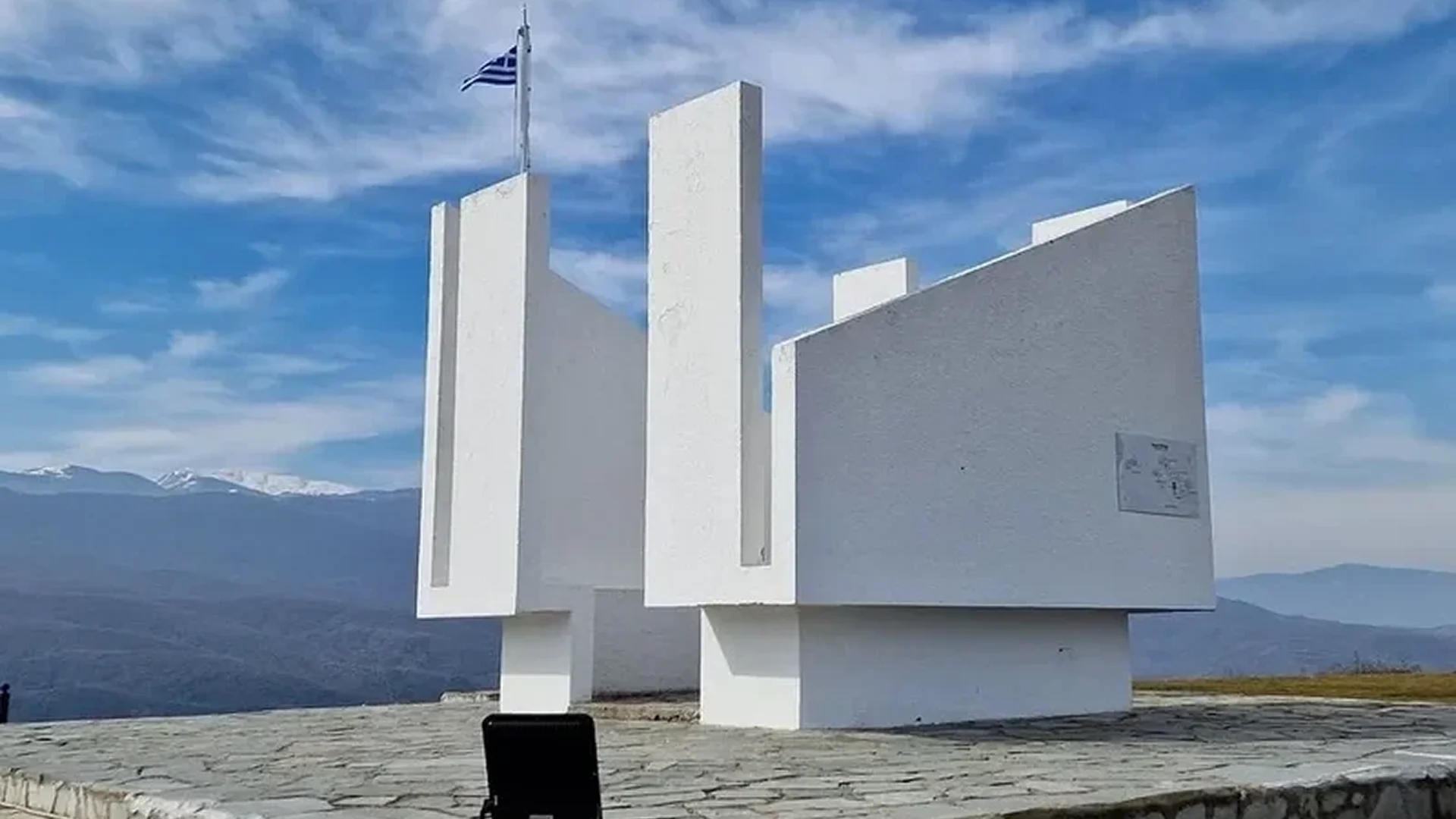 Modern white monument with three tall vertical pillars and a flag against a blue sky.