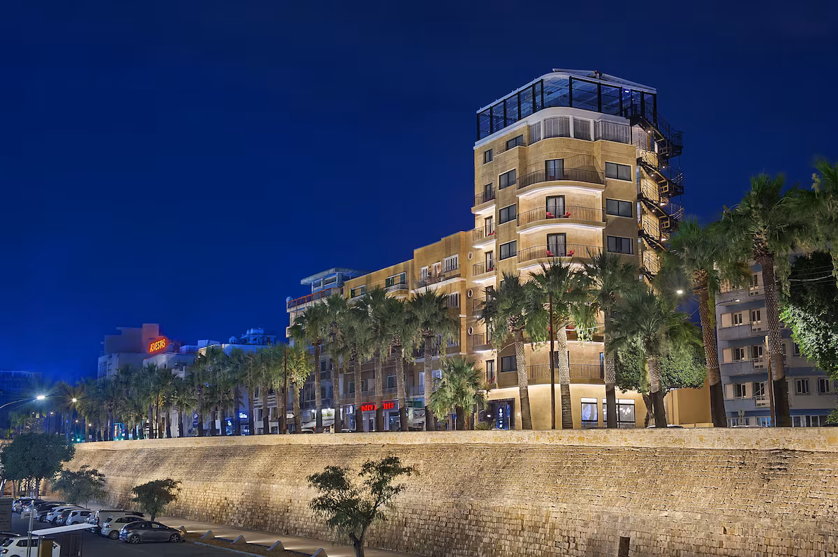 Night view of a lit-up building and palm trees along a waterfront.