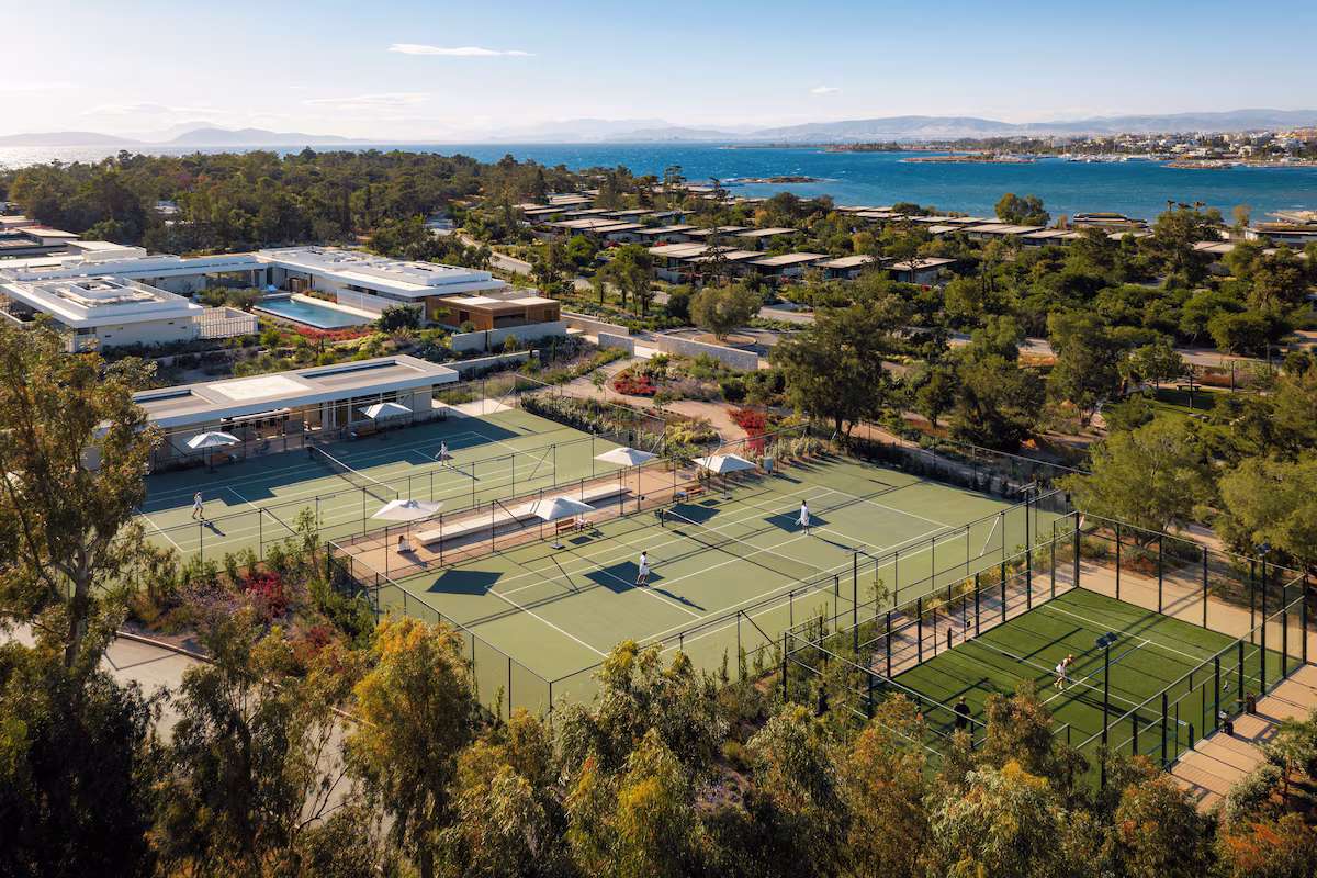 Aerial view of tennis courts near a coastal area with buildings and greenery.