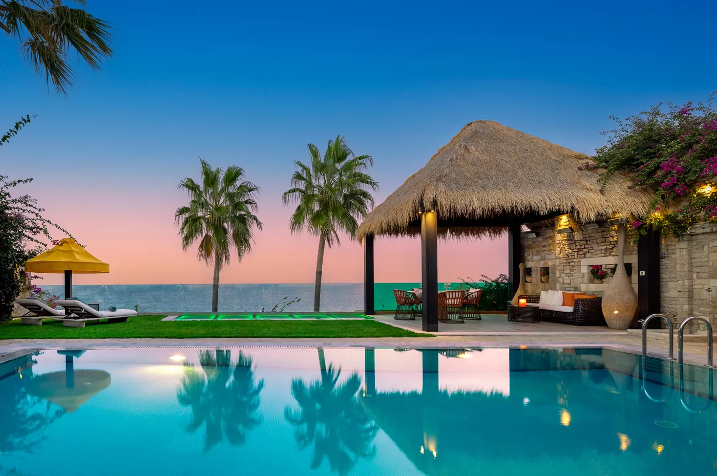 Tropical poolside scene at sunset with palm trees and a thatched-roof bar.