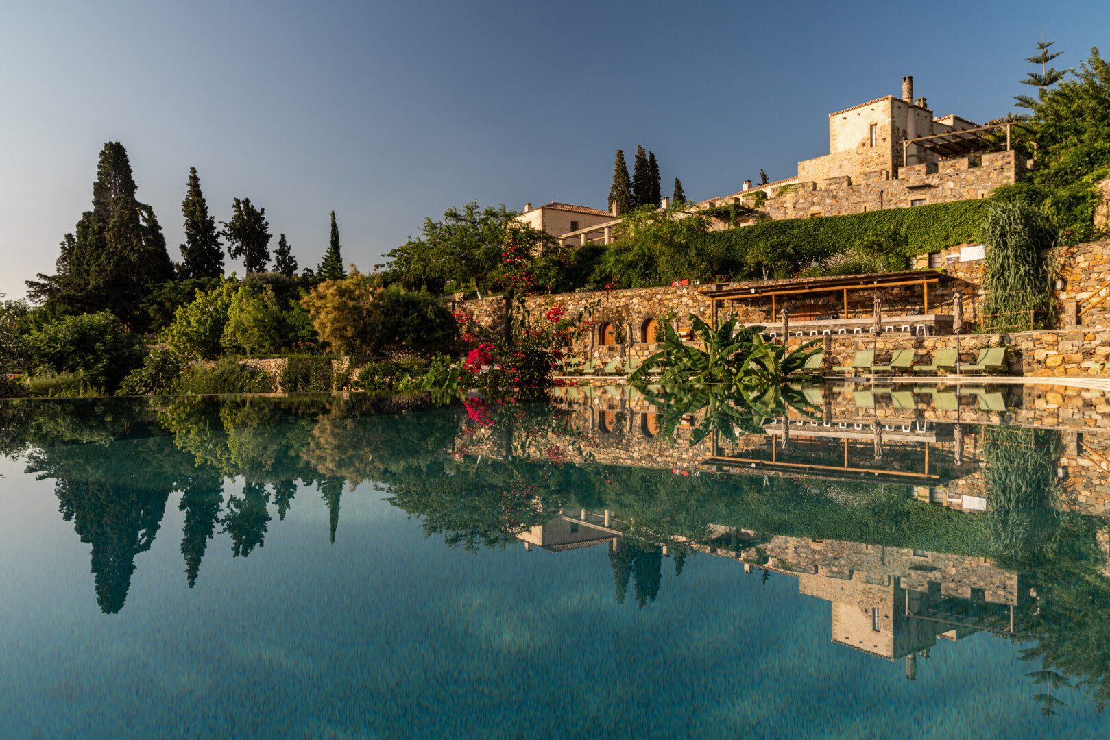 Peaceful water reflecting a sunny hillside with greenery and buildings.