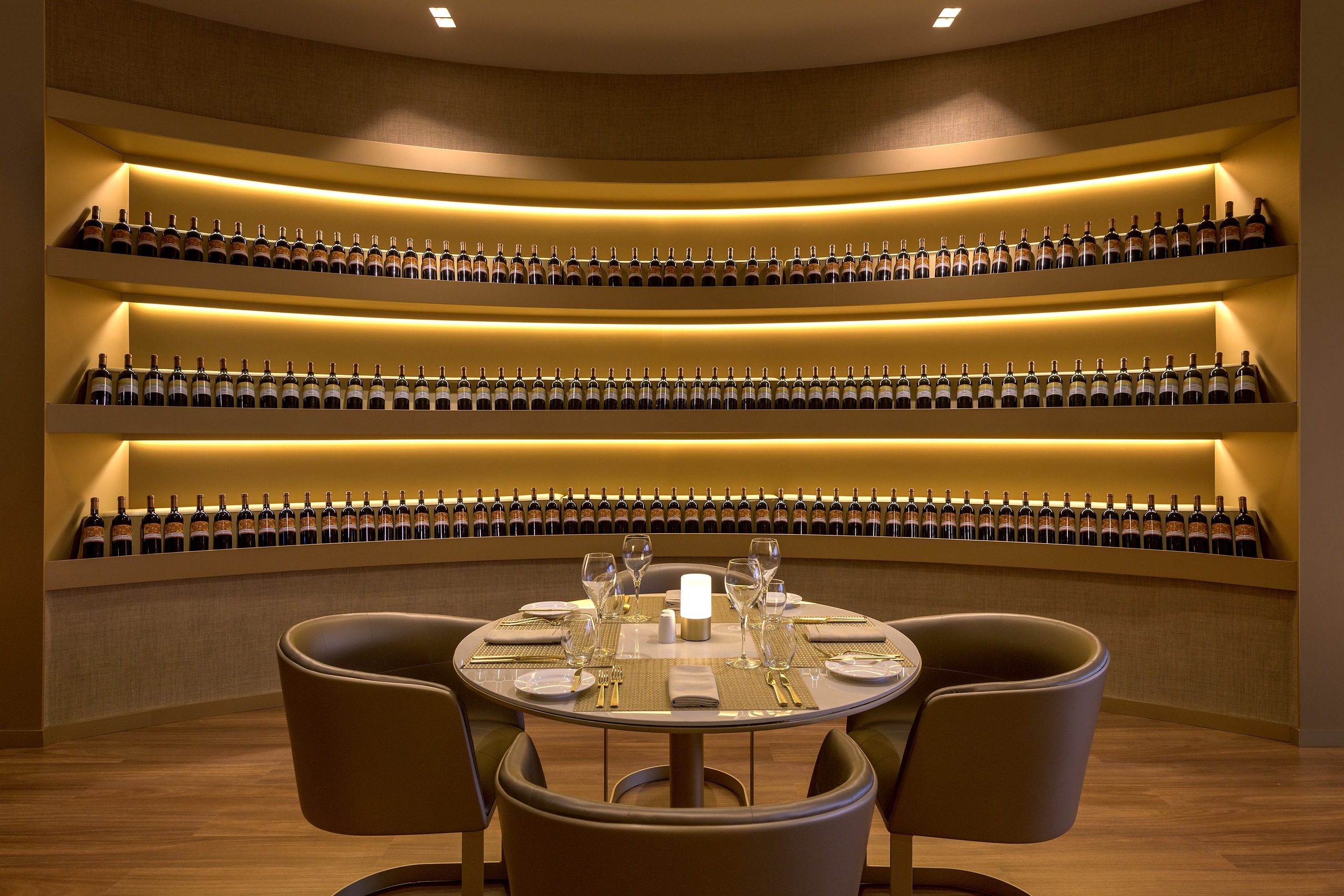 Elegant dining table set with wine glasses against a wall of illuminated wine bottles.