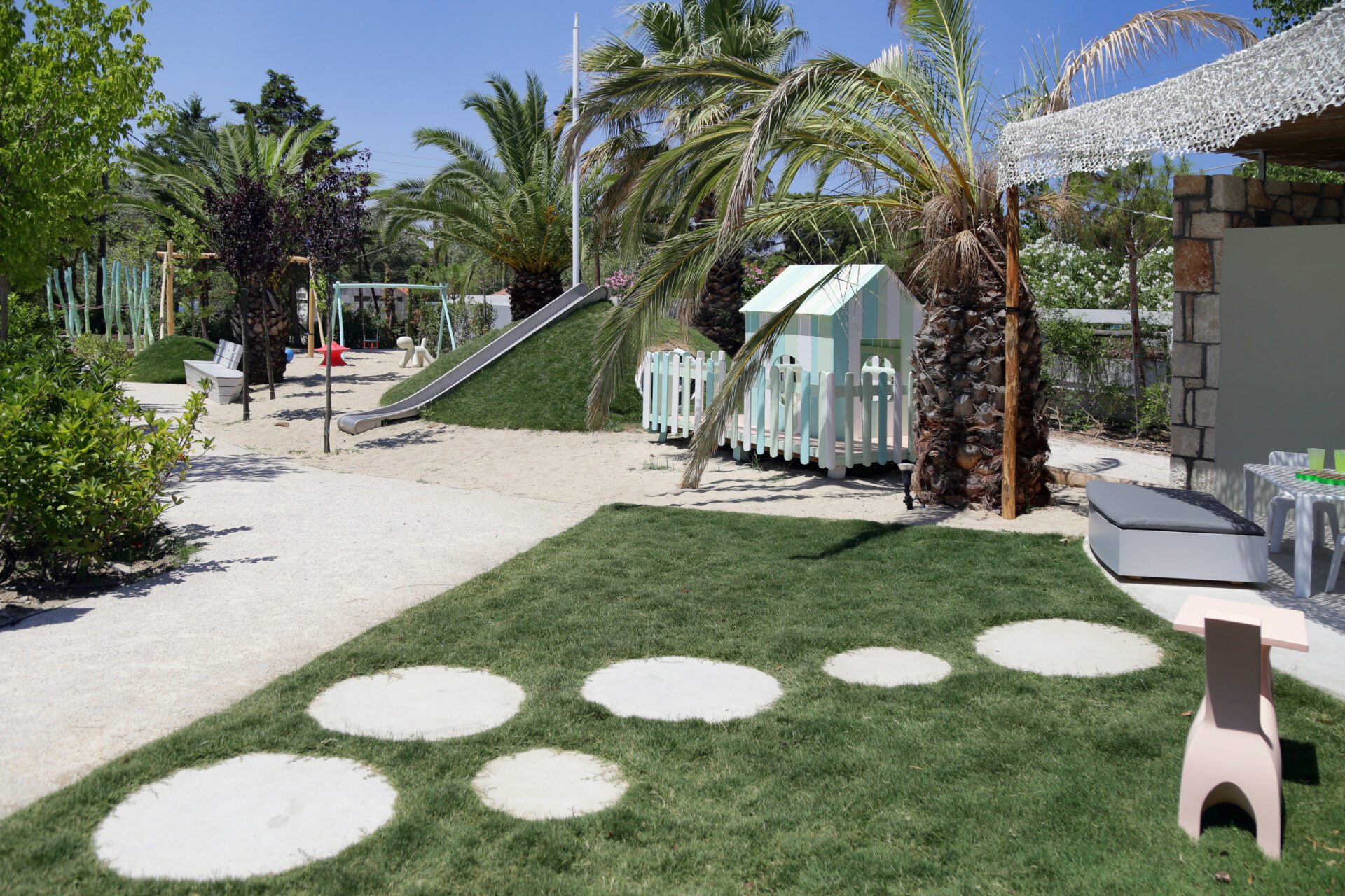 Playground with grass, palm trees, and a slide under a sunny sky.