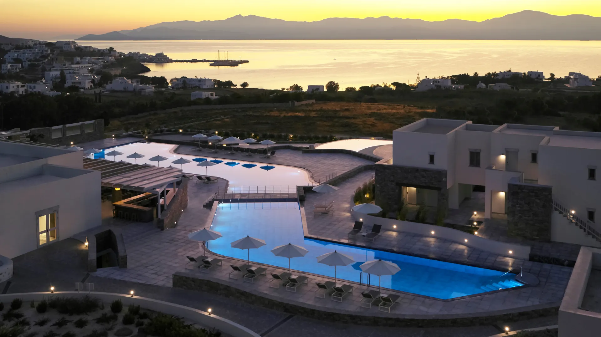 A serene poolside scene at sunset with umbrellas and distant mountains.
