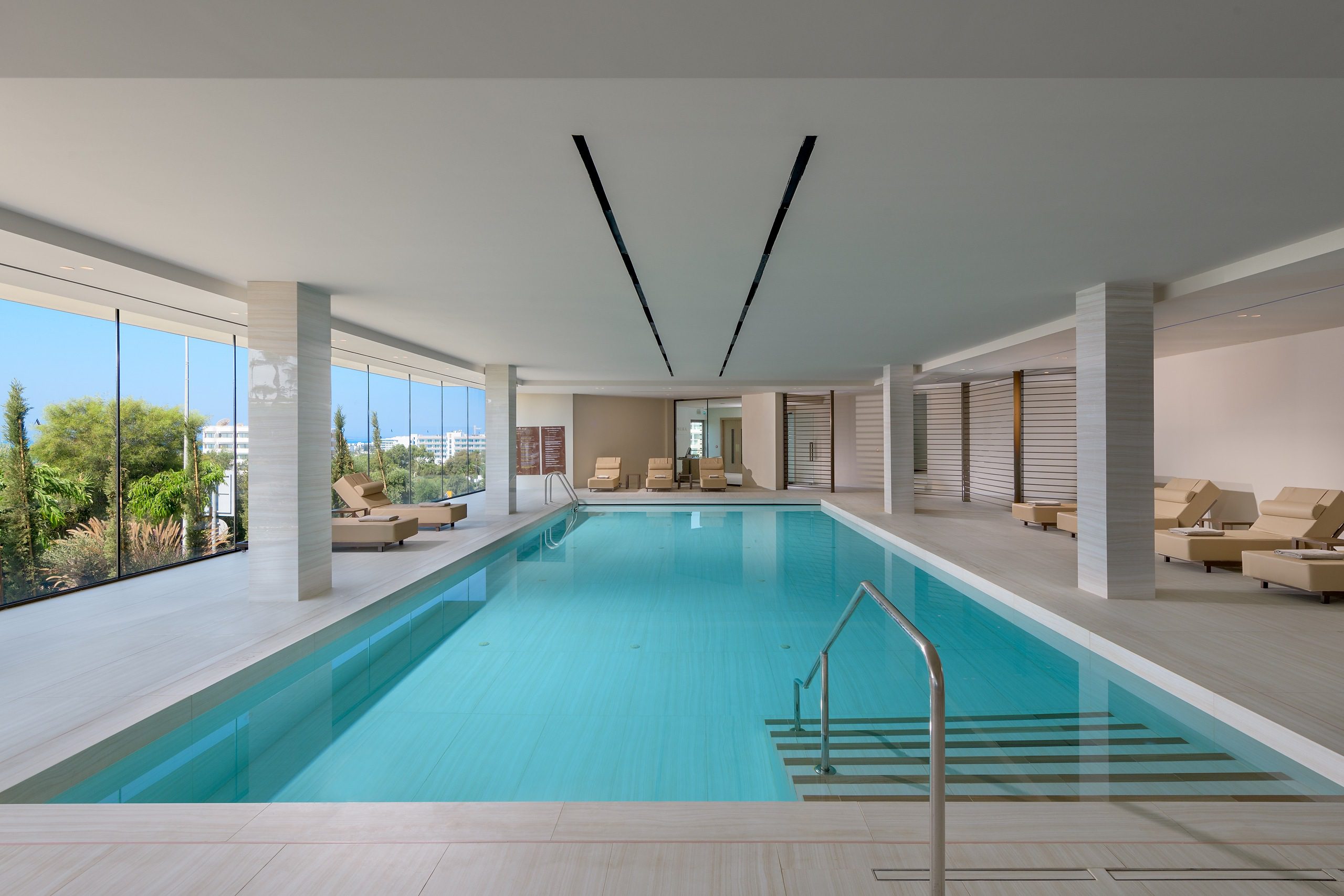 Modern indoor swimming pool with large windows and lounge area.