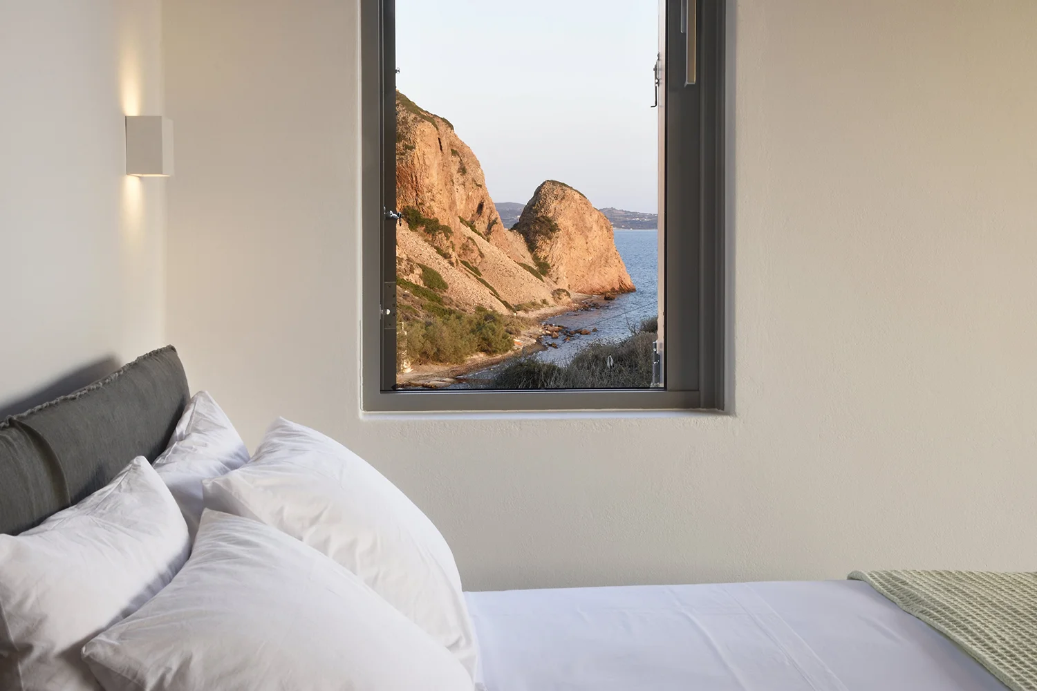 Cozy bed with pillows near a window showing rocky cliffs and ocean.