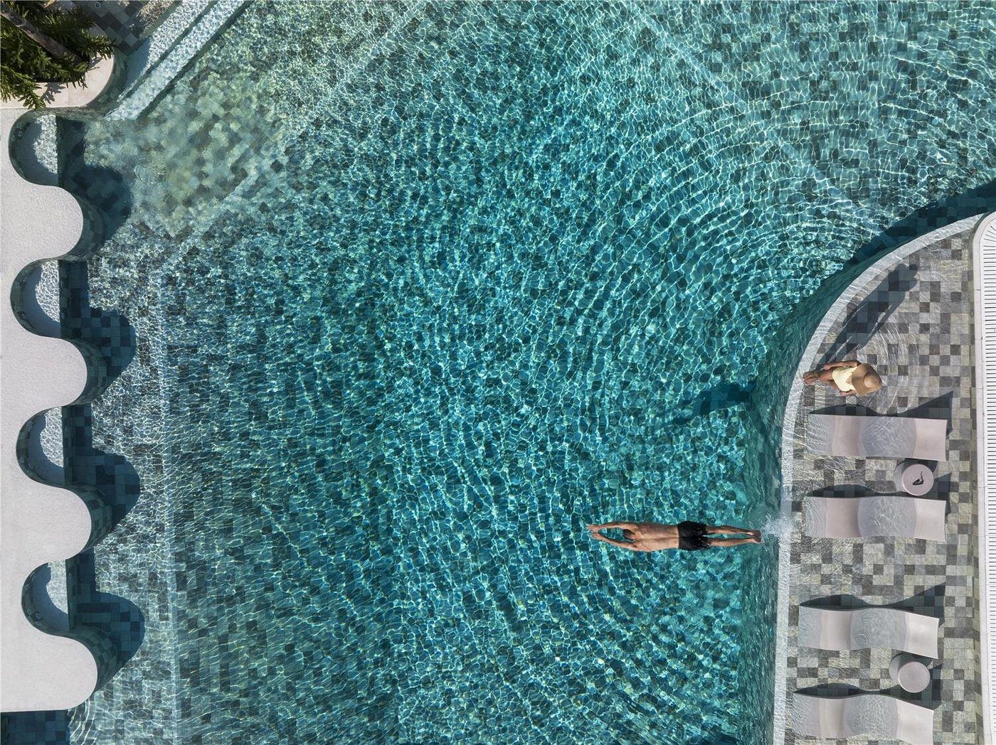 A person floating in a clear blue pool surrounded by lounge chairs.