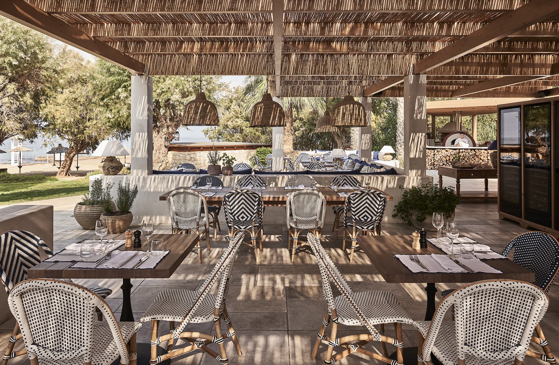 Rustic outdoor dining area with wooden tables and wicker chairs under a thatched roof.