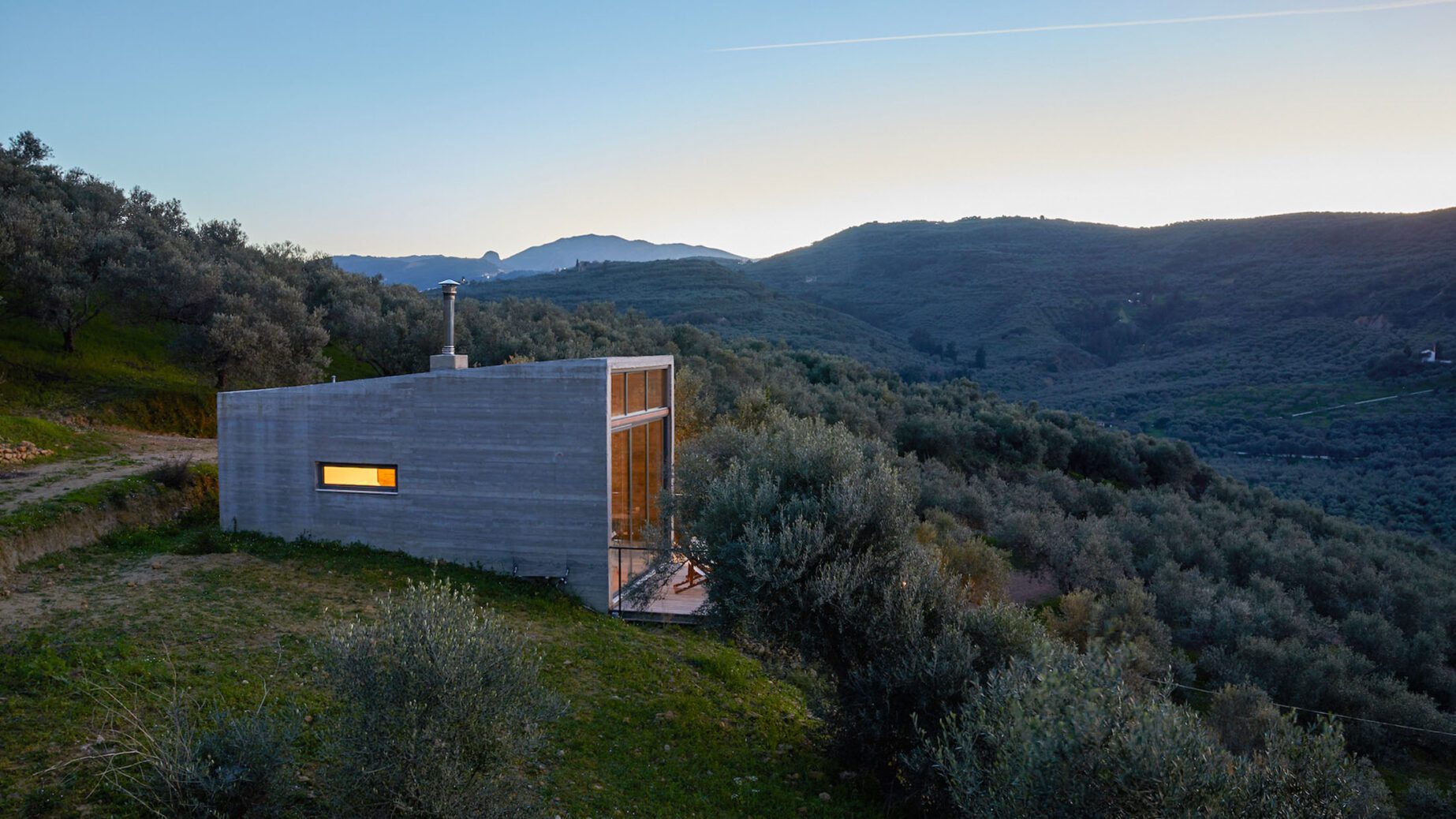 Modern concrete house nestled in a hilly, natural landscape at dusk.