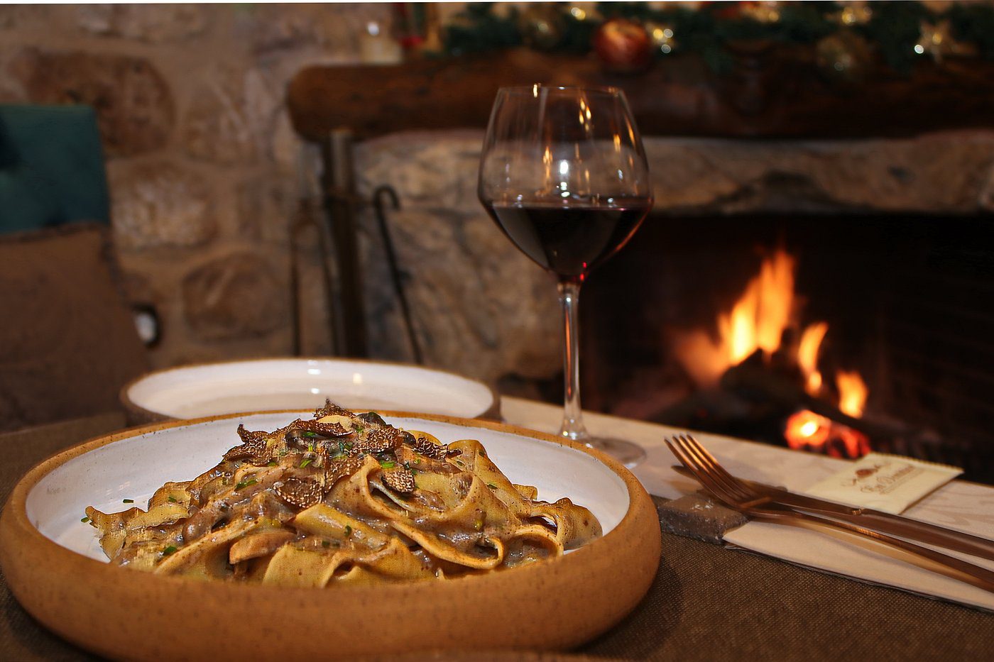 A bowl of pasta with mushrooms and a glass of red wine by a cozy fireplace.