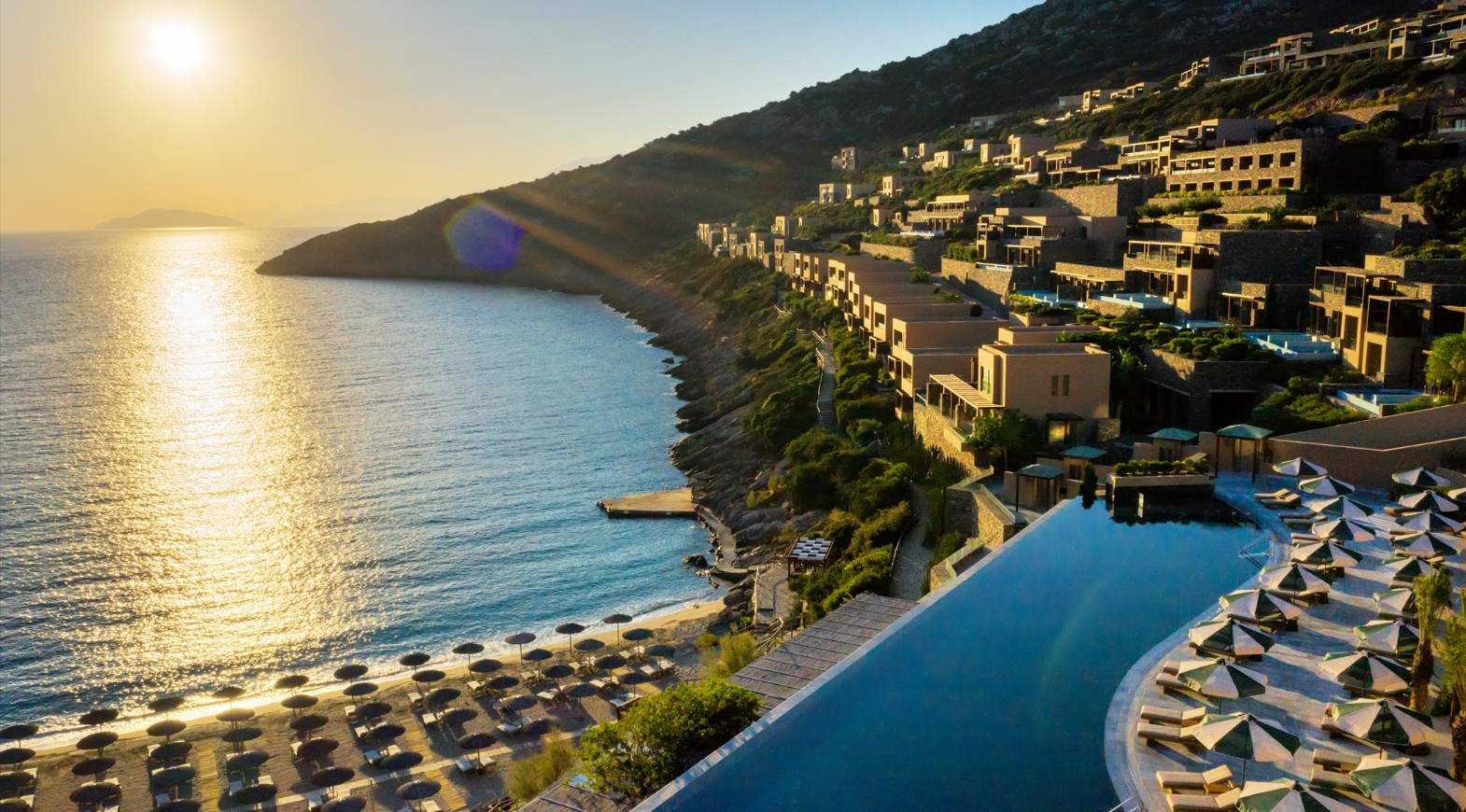 Sunset view of a coastal resort with beach umbrellas and infinity pool.