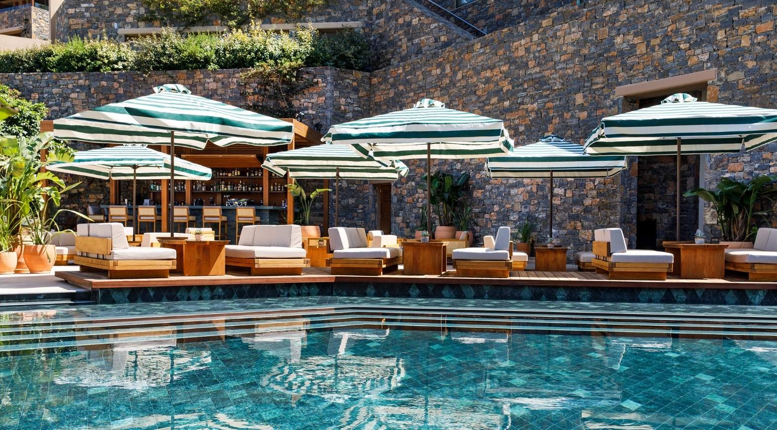 Luxurious poolside lounge area with sunbeds and umbrellas against a stone wall.