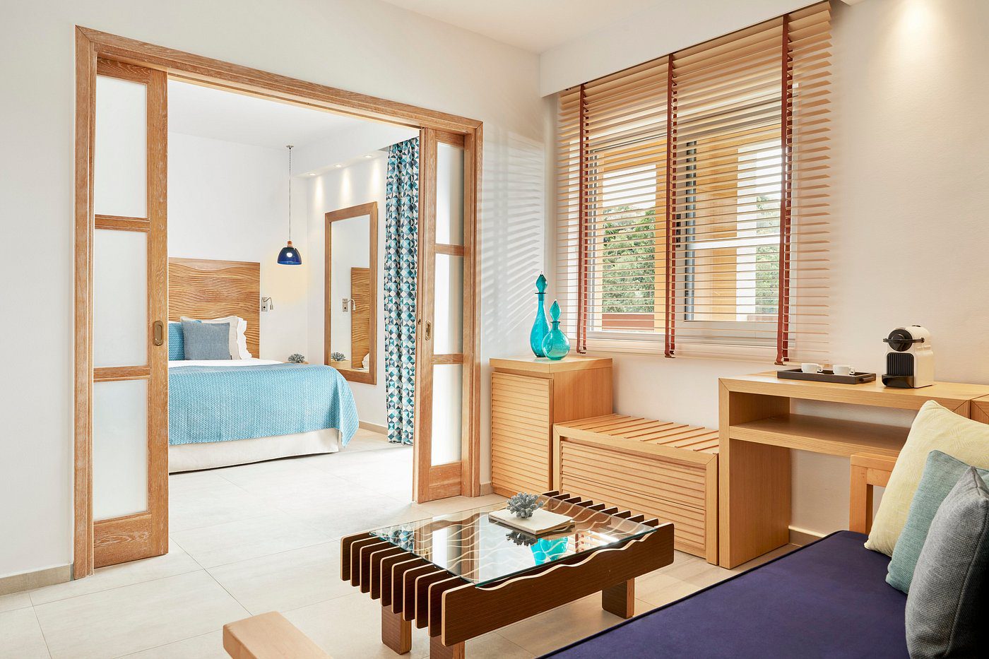 Bright living room with wooden blinds and a view into a cozy bedroom.