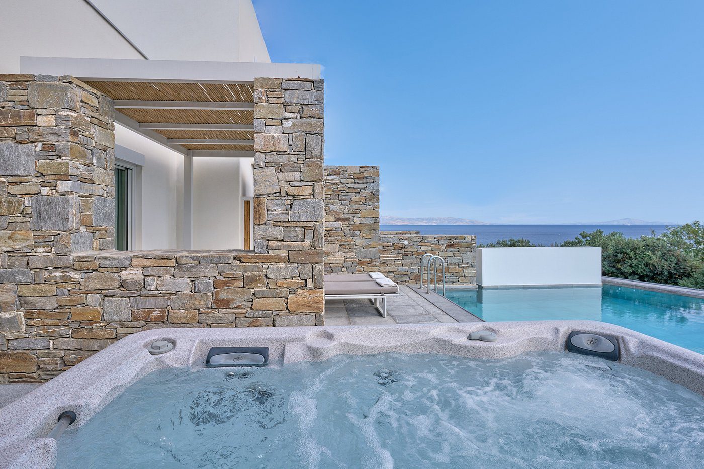 Modern outdoor jacuzzi with stone walls and ocean view under clear blue sky.