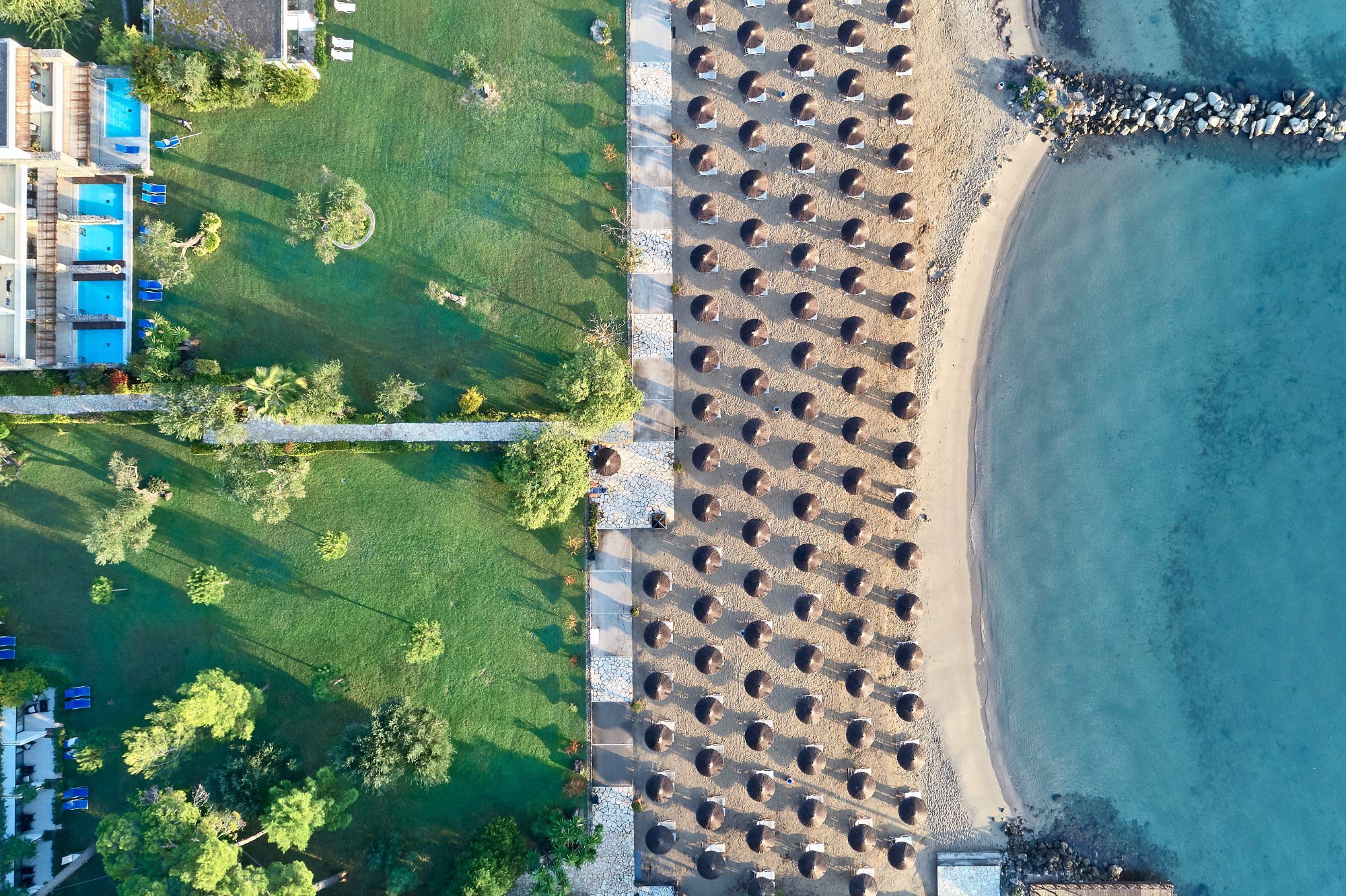 Aerial view of a beach with sun loungers and umbrellas beside a green park.