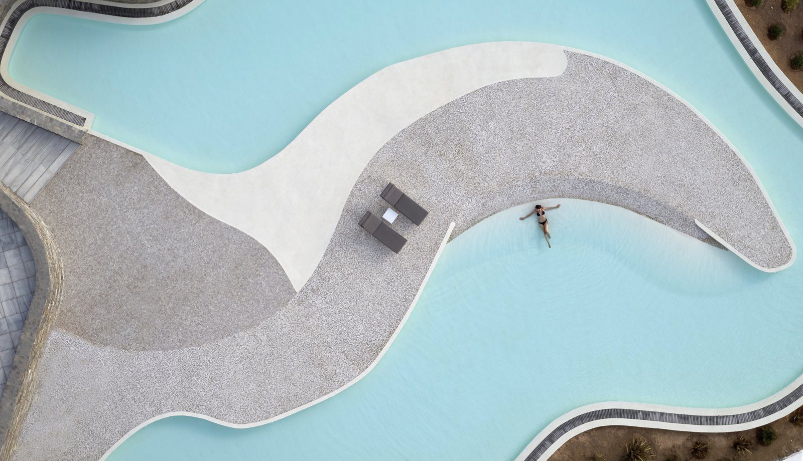 A person swimming near a curved poolside seating area with a table and stools.