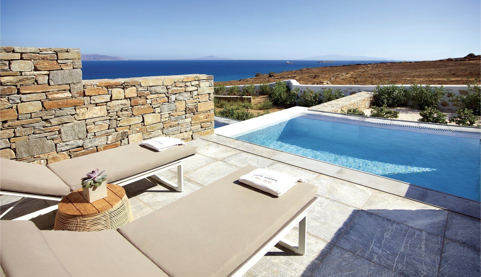Sunlit poolside lounge chairs overlooking the sea and rocky landscape.