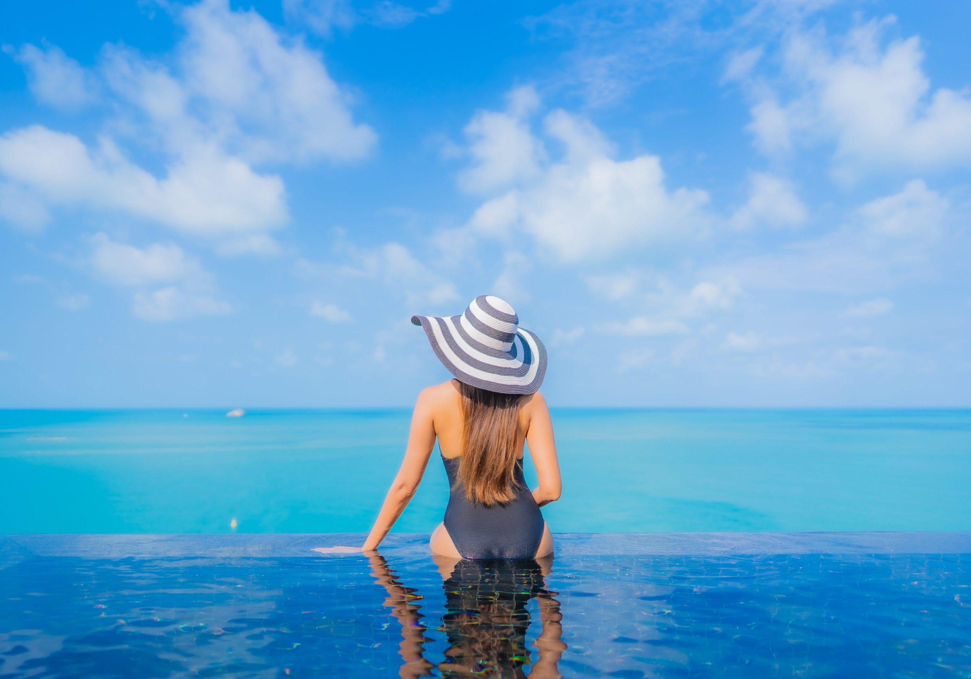 Woman relaxing by an oceanfront pool with a wide sea view, enjoying a peaceful wellness moment under clear blue skies.